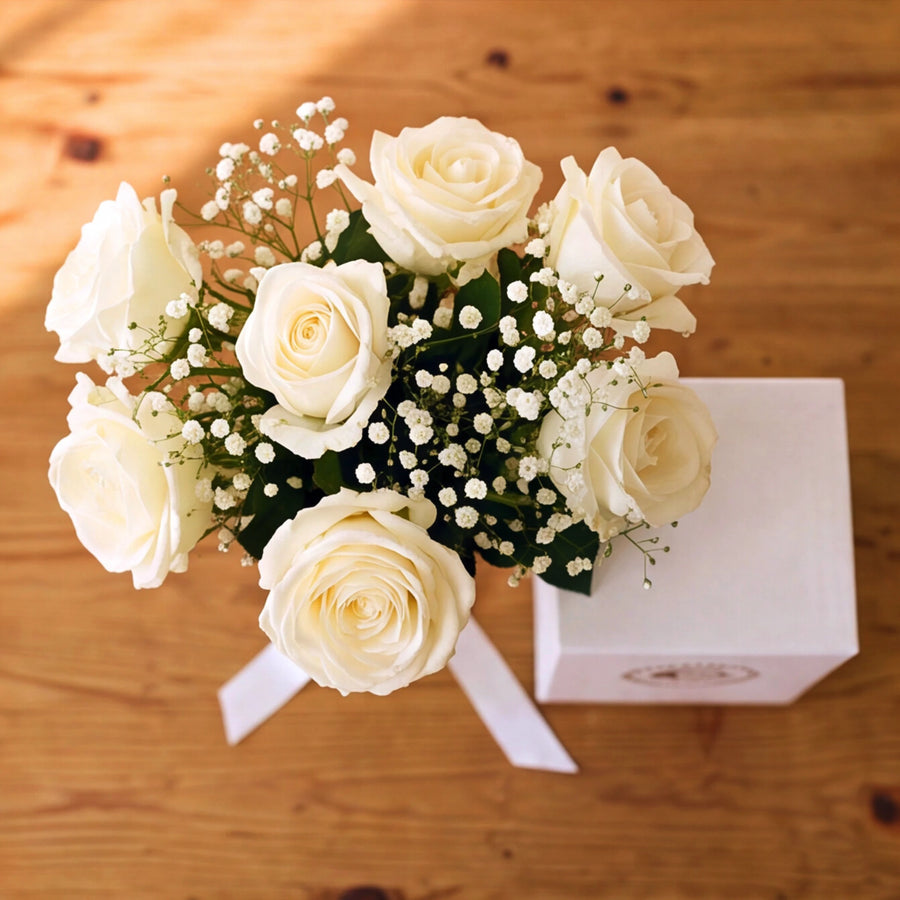 A flower arrangement of white roses and baby's breath with green leaves, tied with a white ribbon, placed on a wooden surface next to a white gift box.