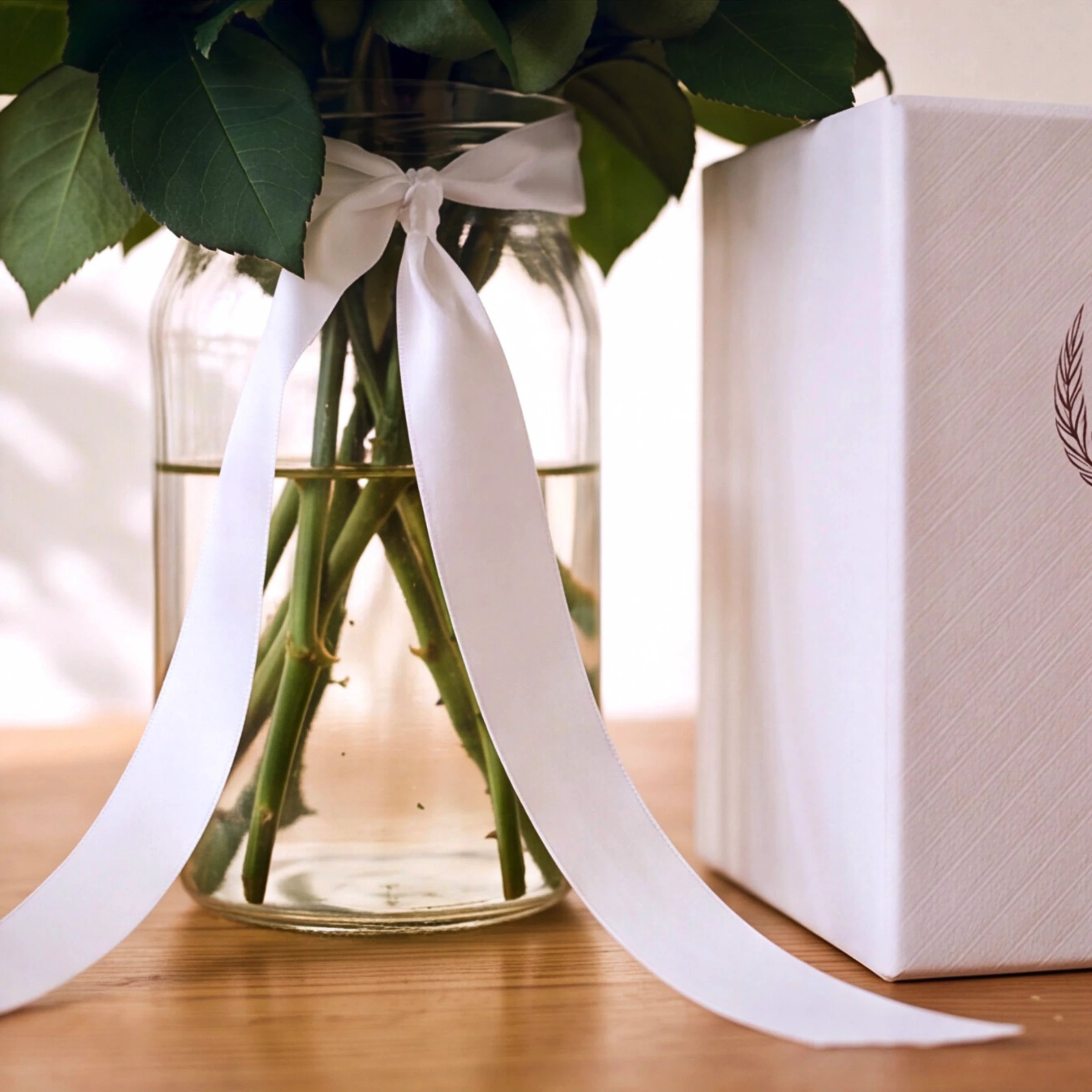 A clear glass jar vase filled with green stems and leaves, tied with a long white ribbon, set on a wooden surface beside a white gift box.