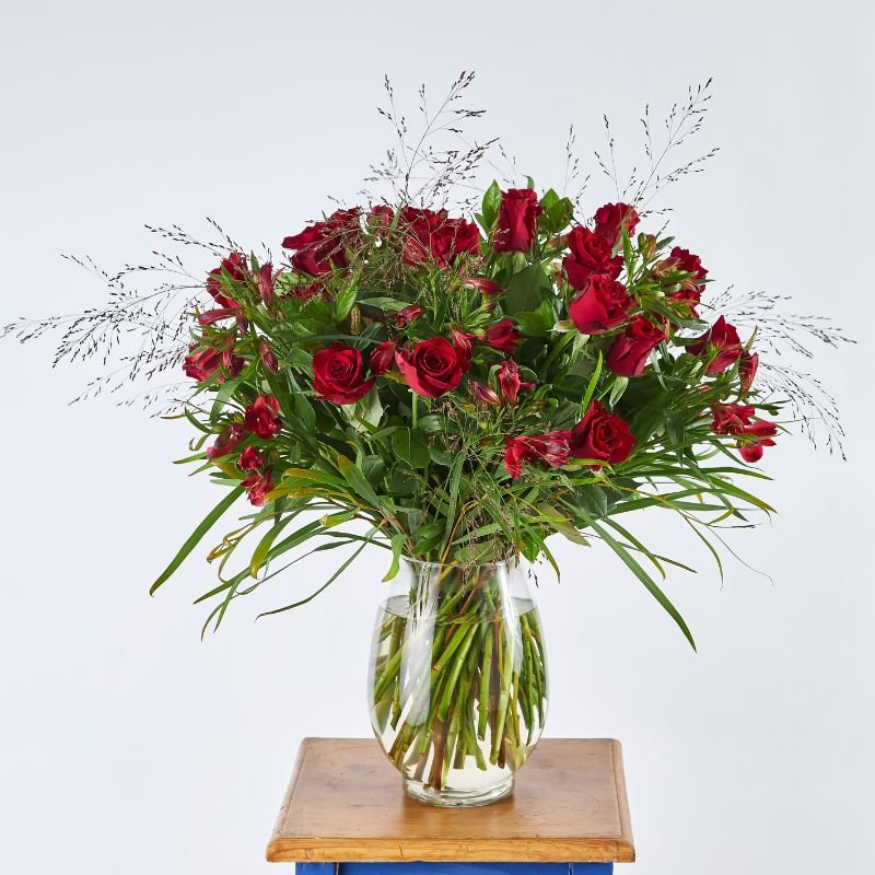 Red Rose of Romance bouquet with two dozen long-stemmed red roses, lush greenery, and filler flowers in a clear glass vase on a wooden table.