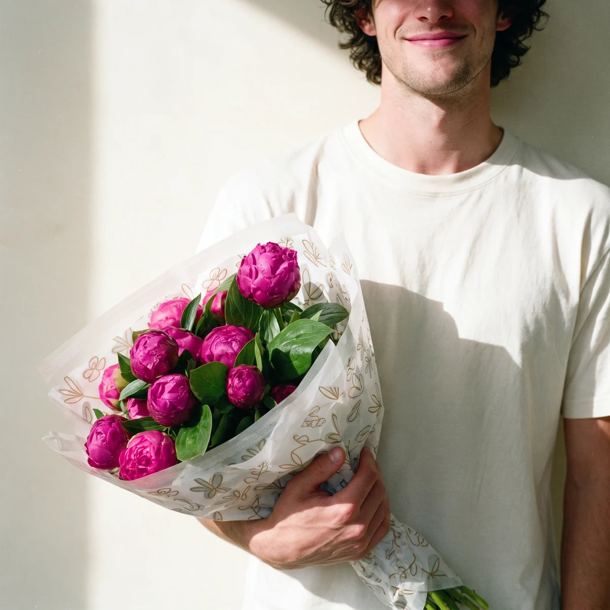 Smiling man with curly dark hair in a white t-shirt holding a large bouquet of twenty magenta peonies against a cream wall, Fabulous Flowers South Africa