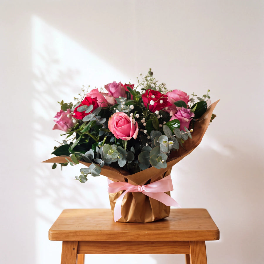 A vibrant flower bouquet featuring pink and red roses, green eucalyptus, and small red baby's breath, wrapped in brown paper with a pink ribbon, displayed on a wooden stool against a white wall with soft shadow patterns.