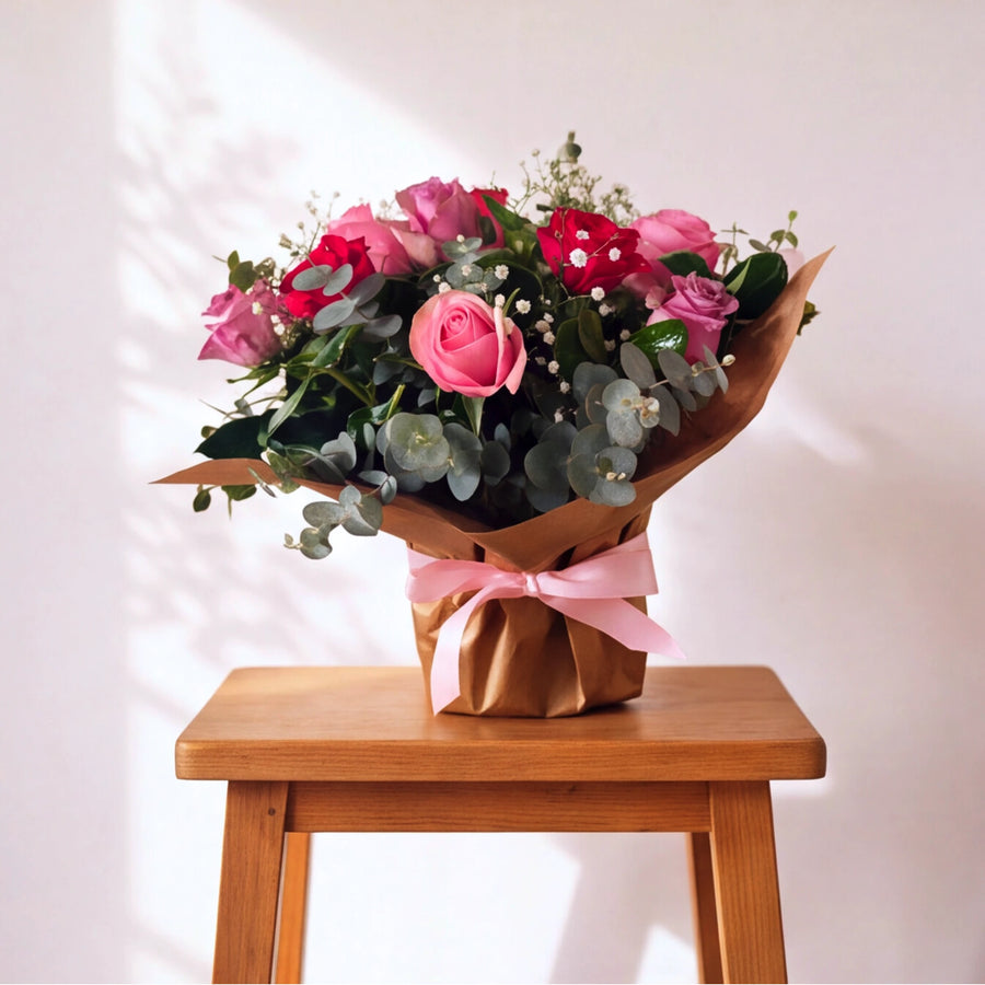 A bouquet of pink and red roses with green eucalyptus leaves and small white filler flowers, wrapped in brown paper with a pink ribbon, displayed on a wooden stool against a light wall with soft shadows.