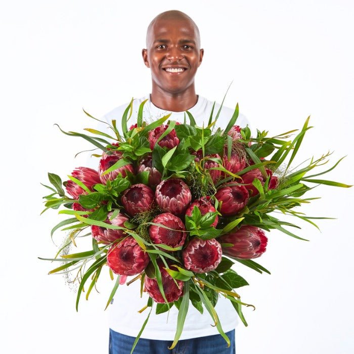 Royal Protea Flower Bouquet – large pink Proteas with lush greenery held by a smiling man in a white shirt.