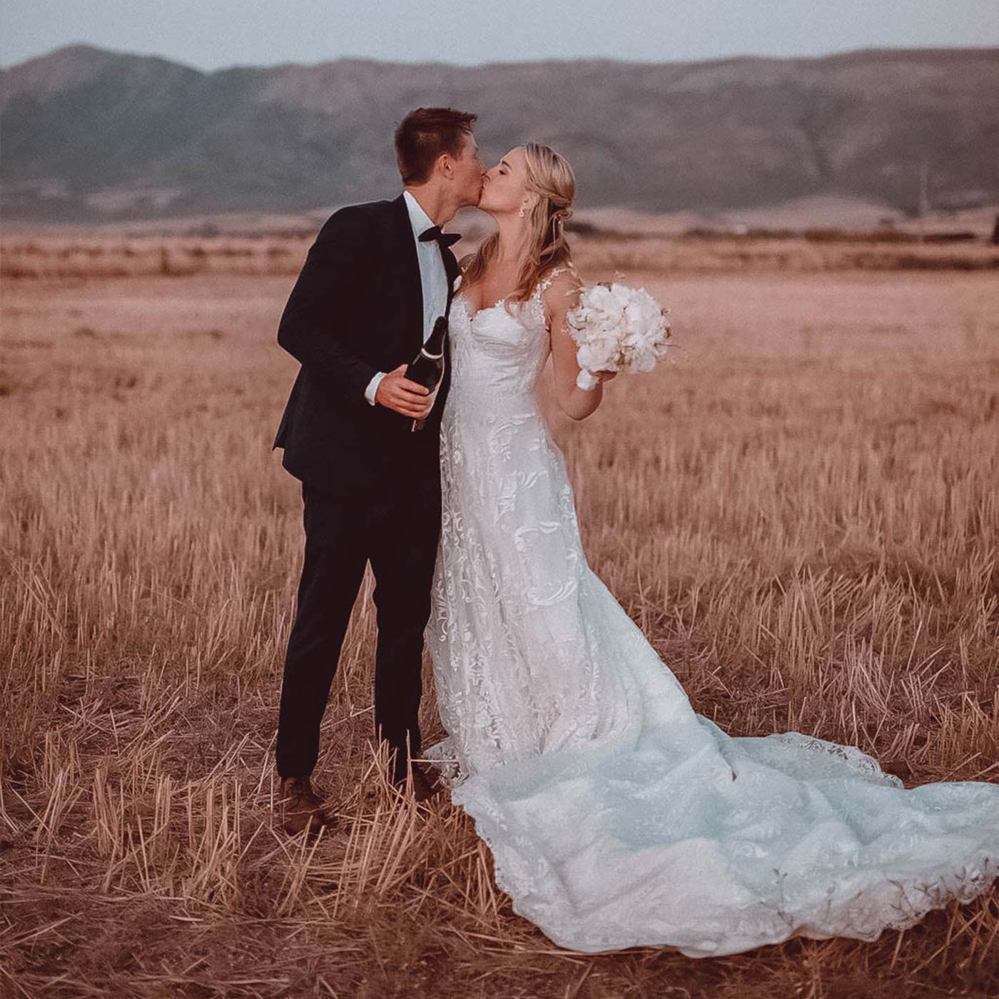 Bride Skye and groom Scott share a kiss in a scenic field, with mountains in the background. Skye holds a bouquet and Scott holds a bottle of champagne. Weddings. Fabulous Flowers and Gifts.