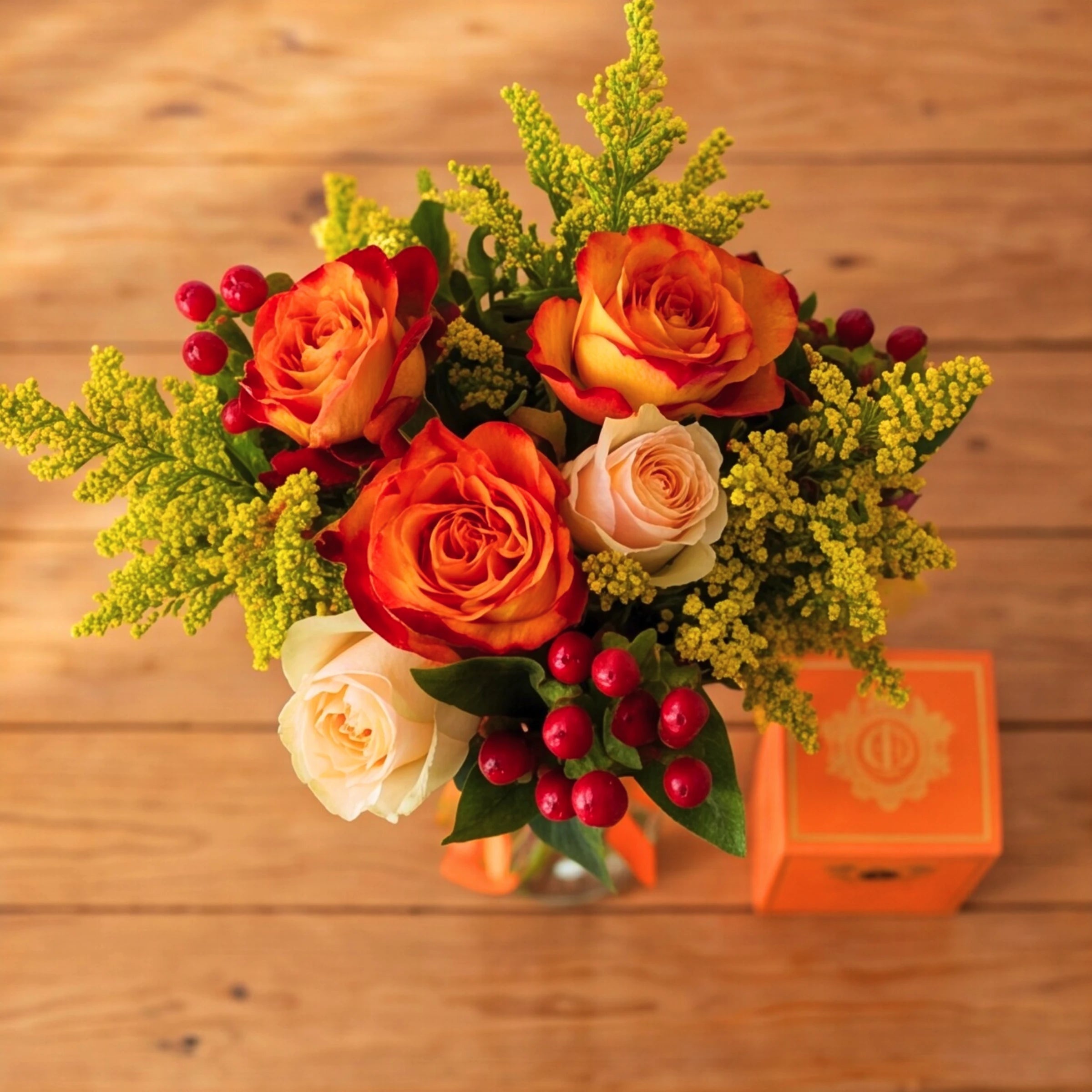 Top down view of the Savanna & Sun posy with roses, berries and solidago next to an orange candle box