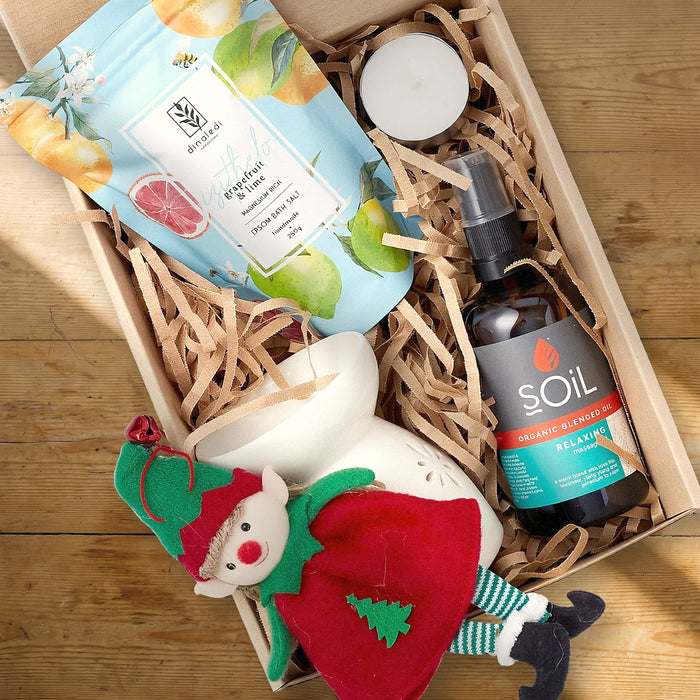 Open kraft gift box from above with a grapefruit and lime Epsom bath salt, a tealight, a white ceramic oil burner, a SOiL Relaxing massage oil and a felt Christmas elf on a sunlit pine table, Fabulous Flowers South Africa