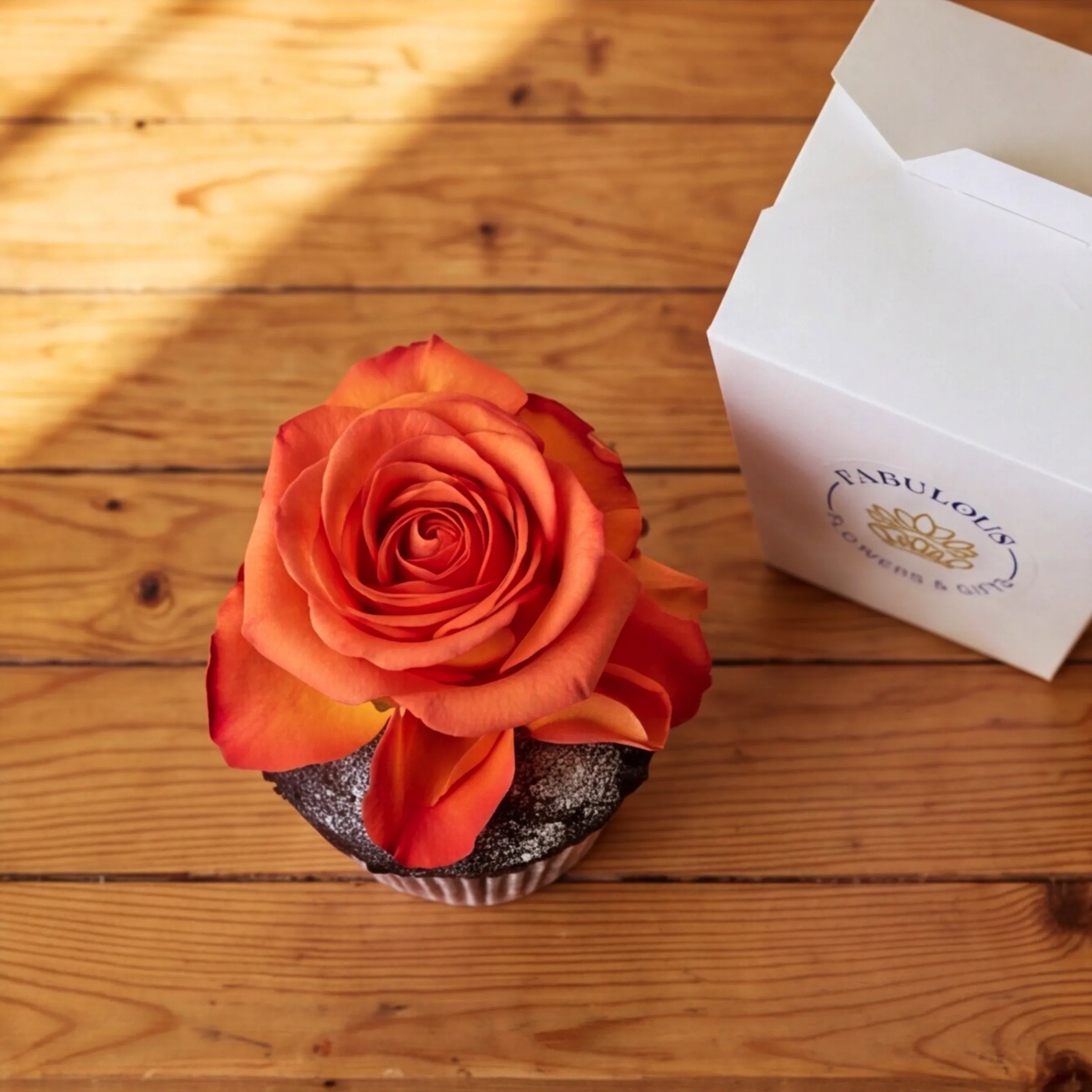 Overhead view of a Fabulous Flowers chocolate cupcake with an orange rose bloom centred on the dark chocolate sponge, white branded box to the side on a rustic wooden surface