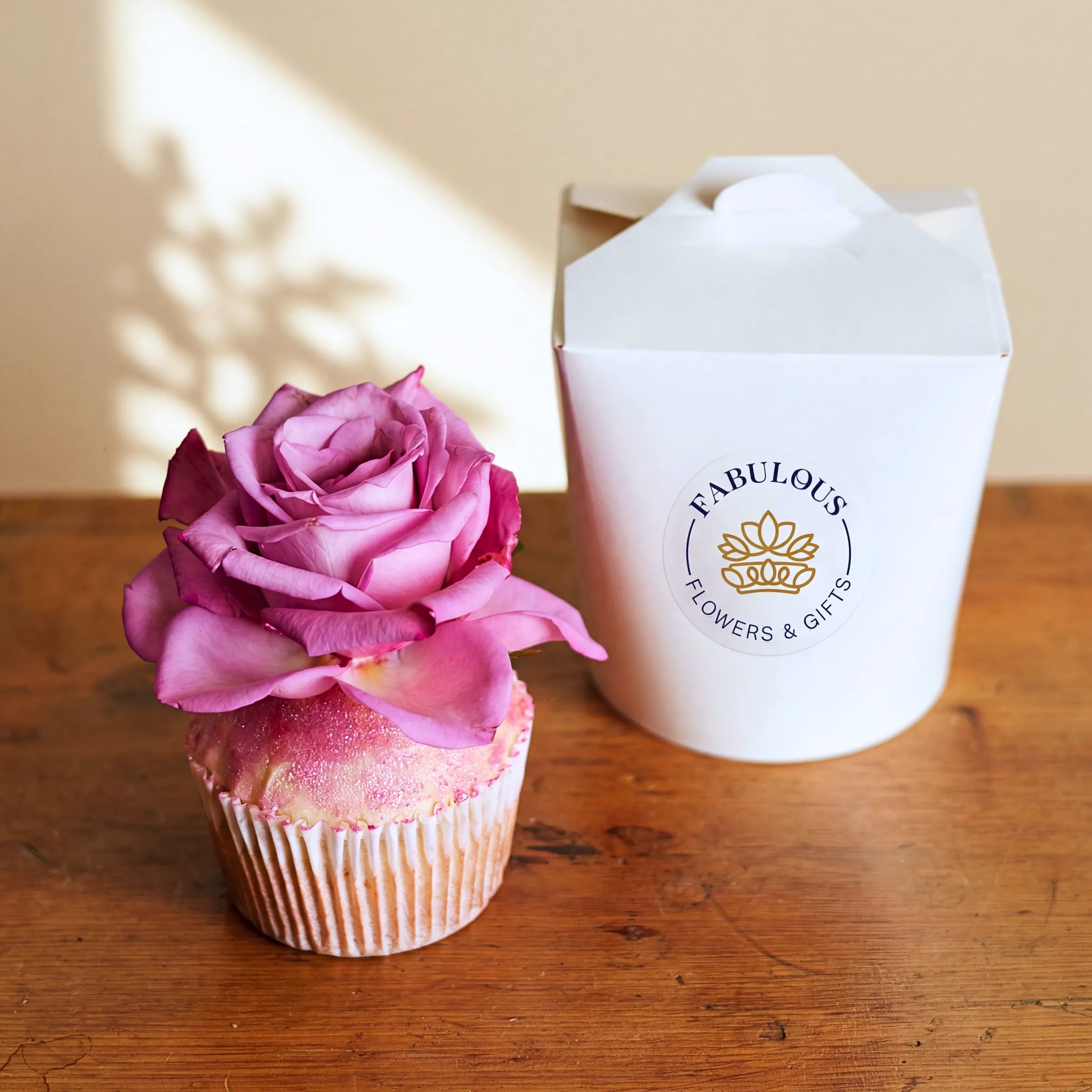 Single Vanilla Cupcake topped with a real pink rose and edible glitter, Fabulous Flowers branded gift box on a wooden table