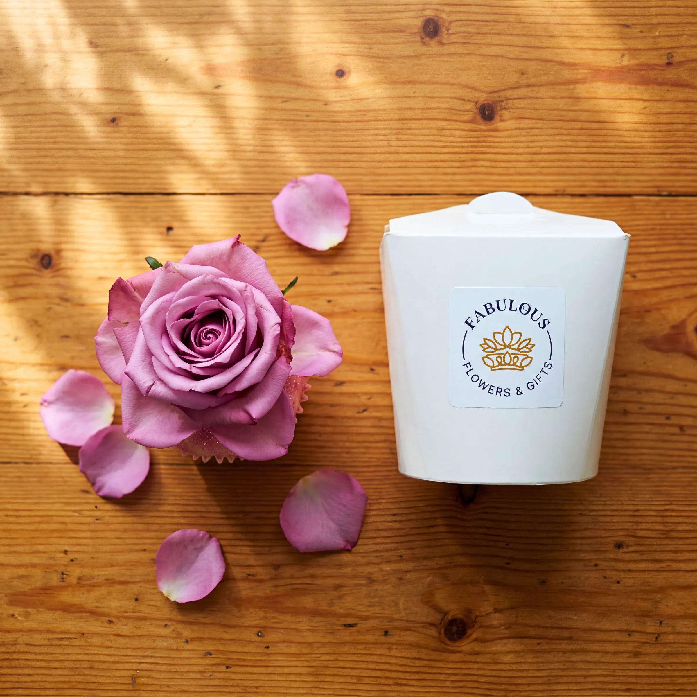 Single Vanilla Cupcake with pink rose, scattered rose petals and Fabulous Flowers gift box on wooden table