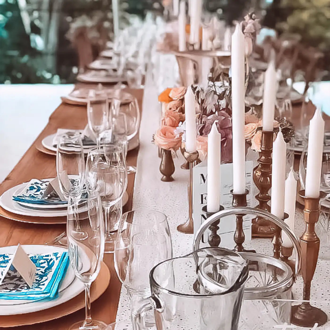 Long banquet table set for an elegant event, adorned with blue and white napkins, glassware, brass candlesticks, and floral centrepieces in soft pink and peach hues. Fabulous Flowers and Gifts.