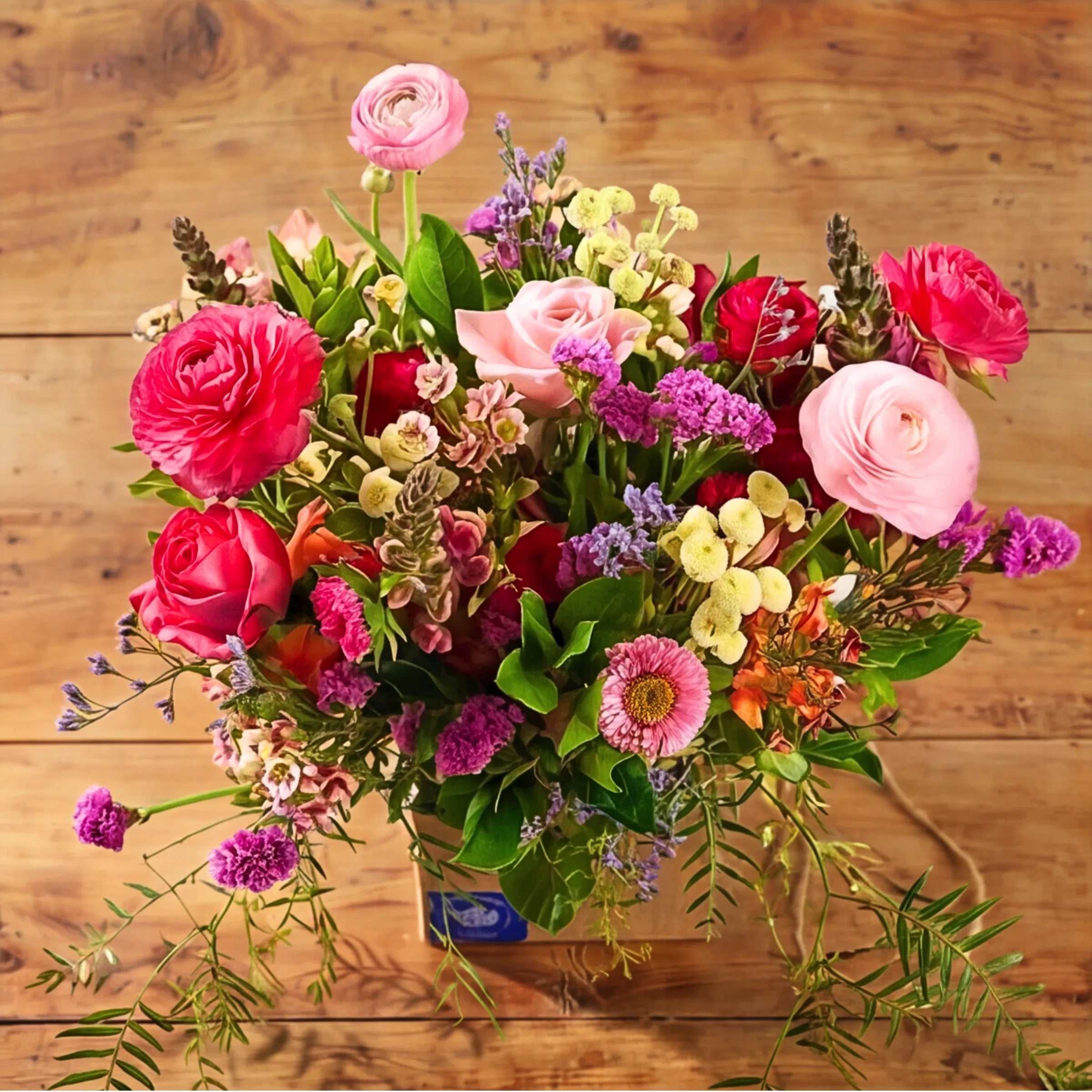 Top-down view of Summer Delight Posy Box showing hot pink and pink ranunculus, hot pink and pink roses, snapdragon spikes, yellow button chrysanthemums, pink aster daisy, purple statice and orange alstroemeria hand-arranged in a kraft posy box, locally grown by Fabulous Flowers South Africa