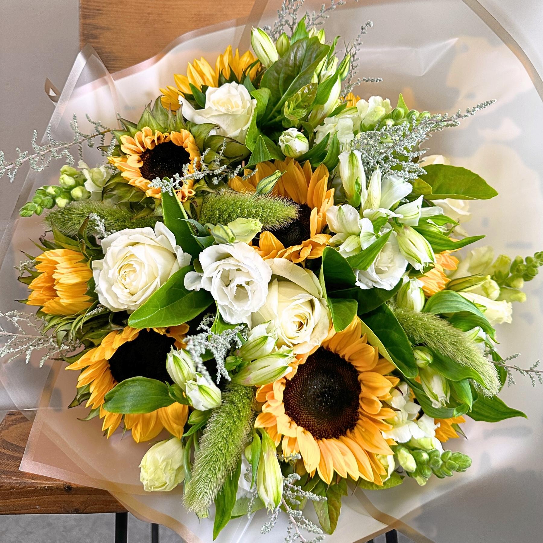 Close-up sunflower bouquet with white roses, lisianthus buds and textured greenery, wrapped in translucent paper