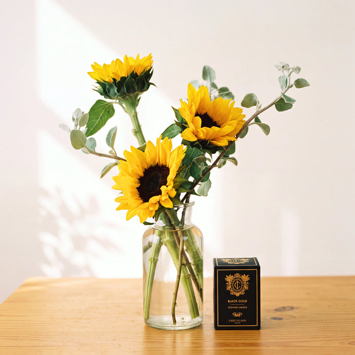 Three yellow sunflowers with green stems arranged in a clear glass jar vase on a wooden table, next to a small black box labeled 'Sunflower' with a gold crest.