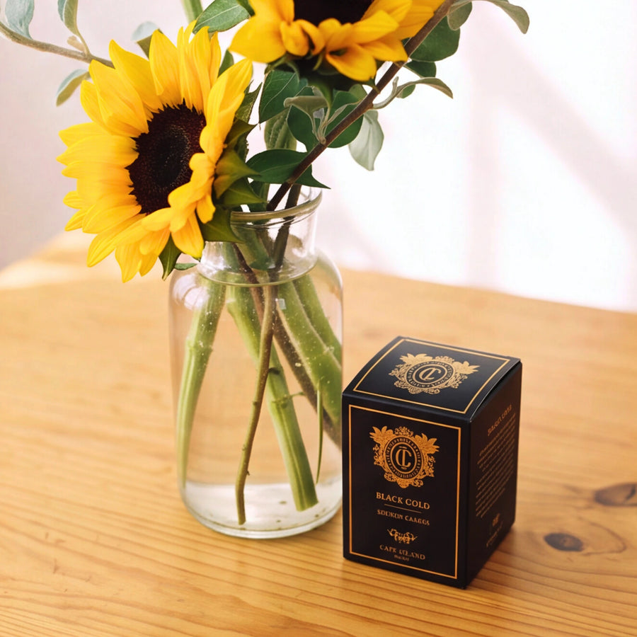 A black and gold box of 'Black Gold' Magic Cards by 'The Art of War' sits on a wooden table next to a glass vase filled with sunflowers.