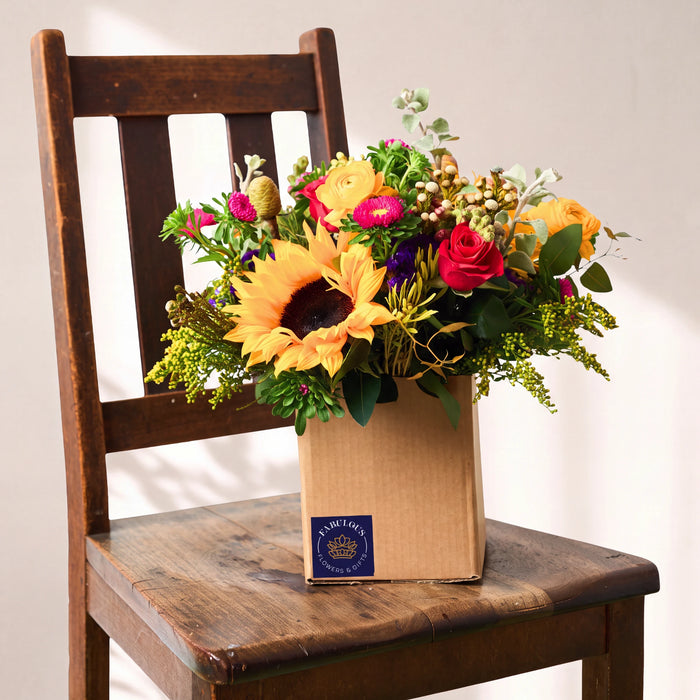A mixed floral arrangement in a brown cardboard box, placed on a dark wooden chair against a light neutral wall. The bouquet includes large yellow sunflowers, red and pink roses, orange blossoms, green foliage, and small filler flowers.