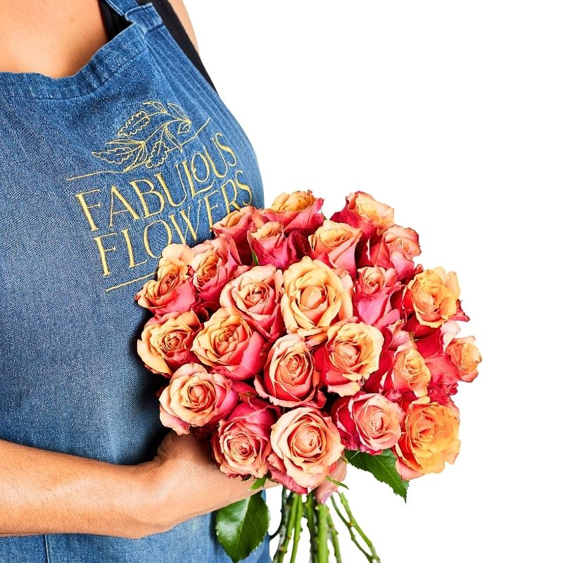 Person holding Sunset Rose Bunch of orange roses in sunset tones, hand-tied bouquet against a blue apron