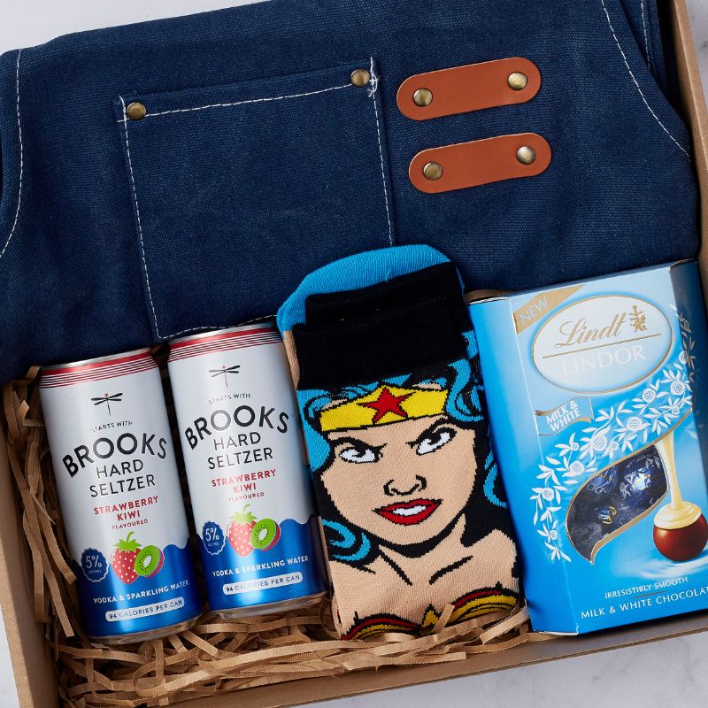 Gift box featuring Brooks Hard Seltzer cans, a blue apron with leather accents, Wonder Woman-themed socks, and Lindt Lindor milk and white chocolate.