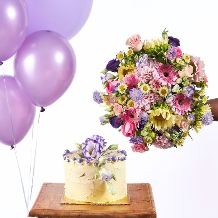Lavish Sweet Celebration gift featuring purple balloons, an assorted flower bouquet, and a yellow cake adorned with purple flowers, set against a white background.