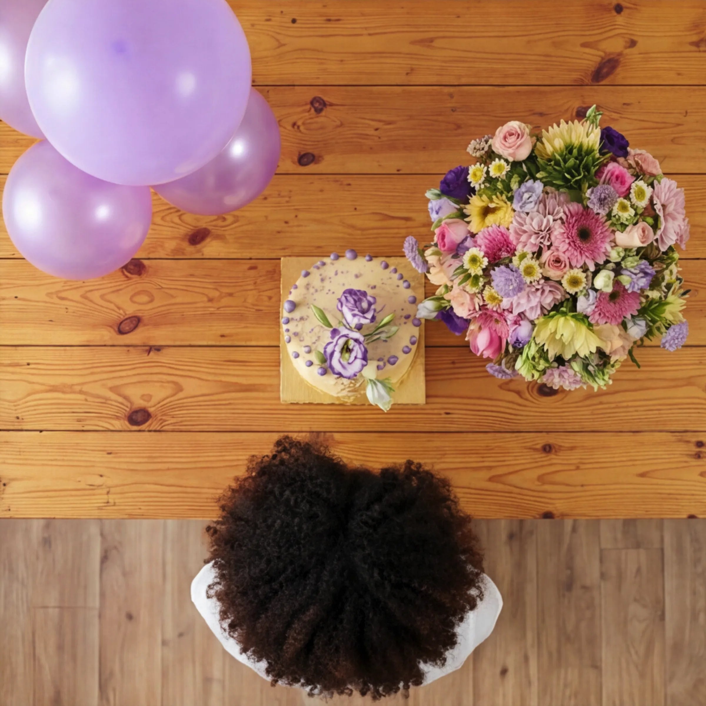 Overhead flat lay of Sweet Celebration with bouquet, buttercream cake and lilac balloons on pine table