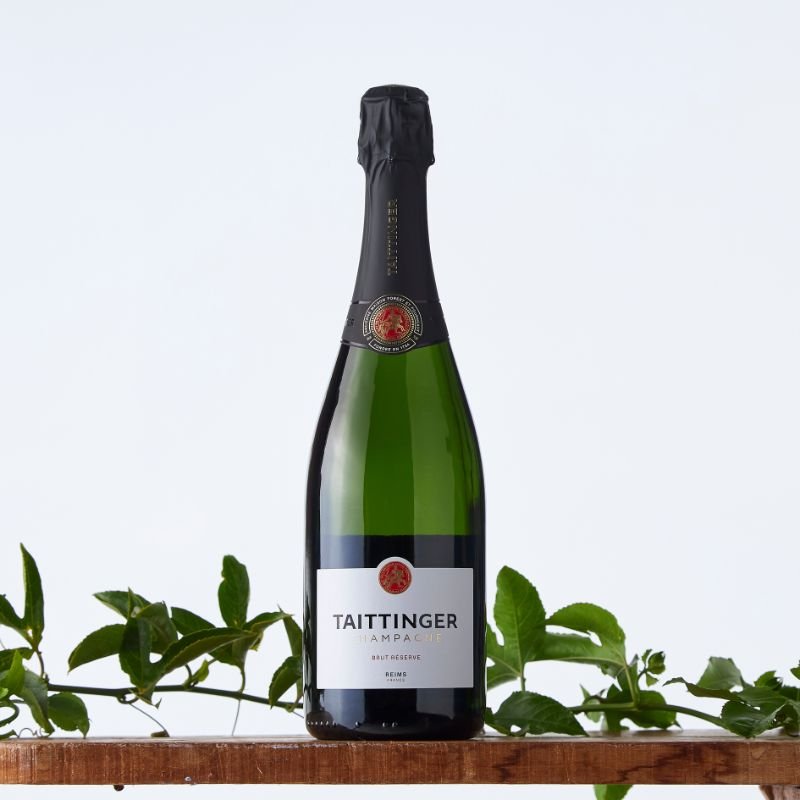 Bottle of Taittinger Brut Réserve champagne displayed on a wooden shelf with green vine leaves against a light background.