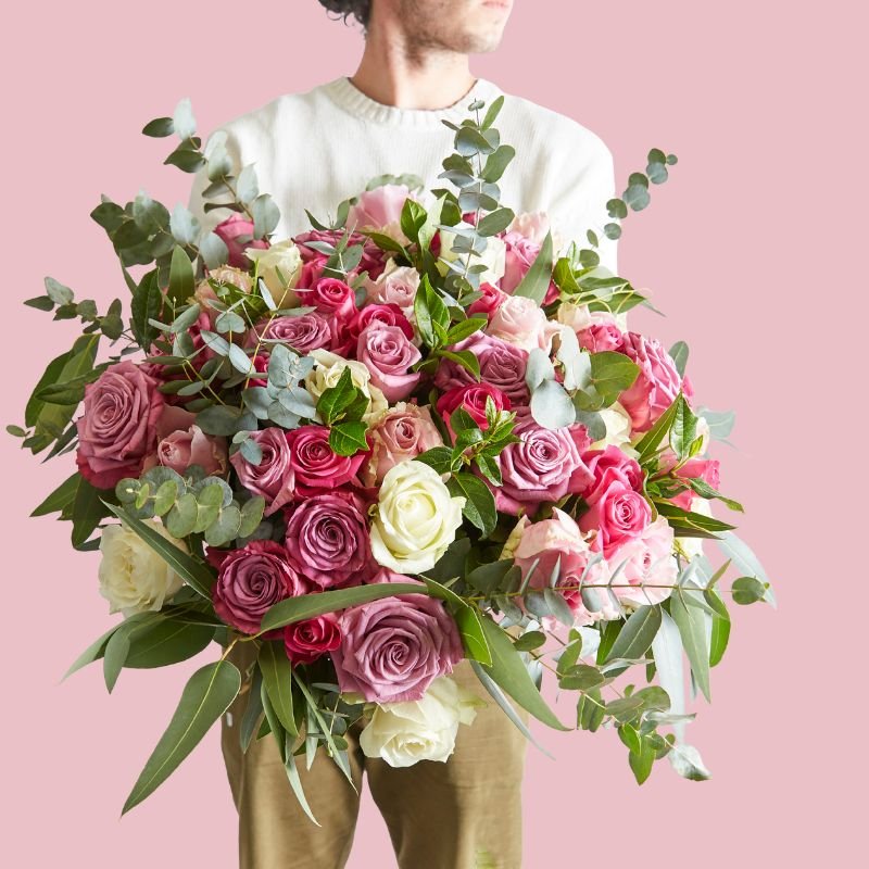 Person holding a large bouquet of pink and white roses with greenery against a pink background