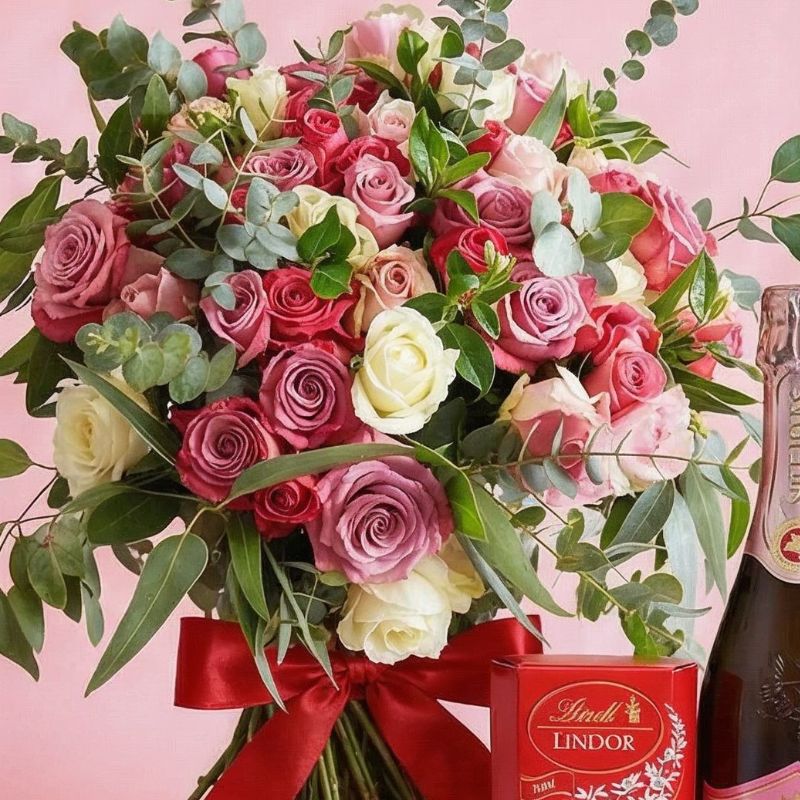 Bouquet of pink and white roses with a Lindor truffles box and bottle of champagne on a pink background