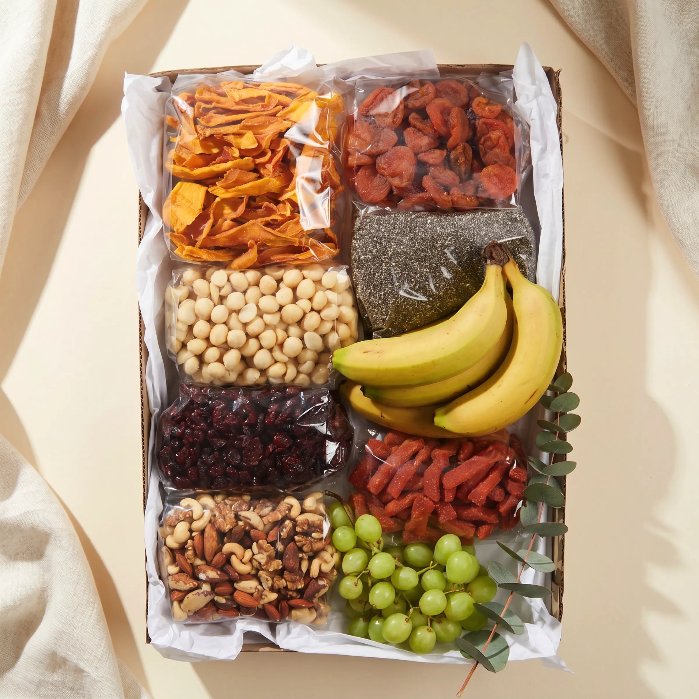 Top-down view of The Healthy Collection by Fabulous Flowers South Africa, eight bags of premium dried fruit, nuts, seeds and fresh fruit hand-packed in a kraft gift box, including dried apricots, sliced mango, meeboos, cranberries, chia seeds, macadamia nuts, mixed nuts, fresh bananas and green grapes