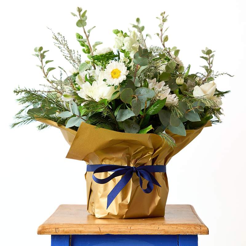Container of flowers wrapped in gold paper with a blue ribbon on a wooden surface.