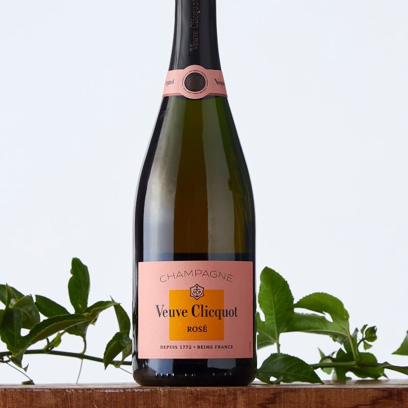 Elegant Veuve Clicquot Rosé champagne bottle with pink foil and label, displayed with greenery on a wooden ledge against a pale background.