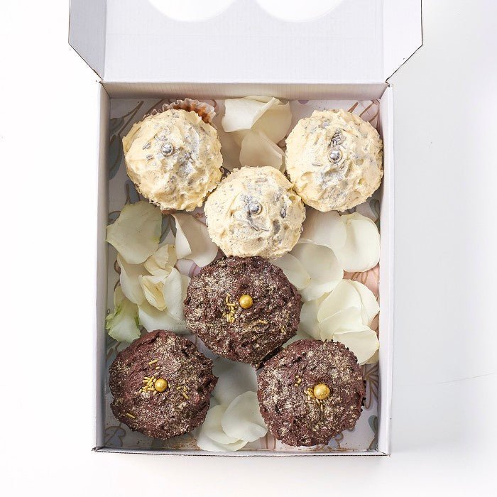 An exquisite box of Where Frosting Meets Fate cupcakes, showcasing a blend of golden-frosted vanilla and rich chocolate cupcakes amidst delicate white petals.