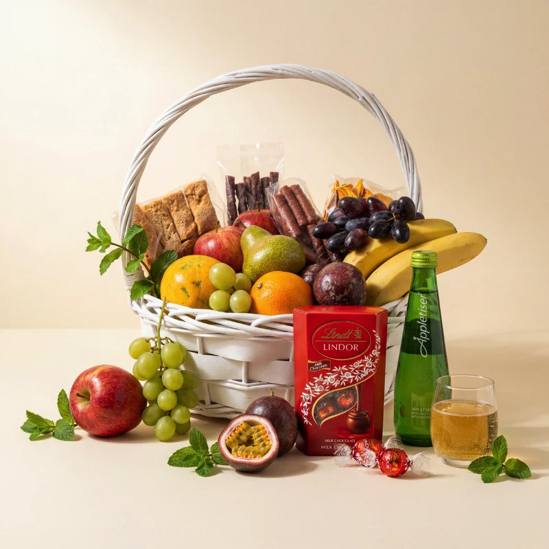 Wholesome Wishes fruit basket with grapes, apples, pear, oranges, bananas, passion fruit, banana bread, biltong sticks, dried mango, Lindt Lindor Milk Chocolate and Appletiser in a white wicker basket