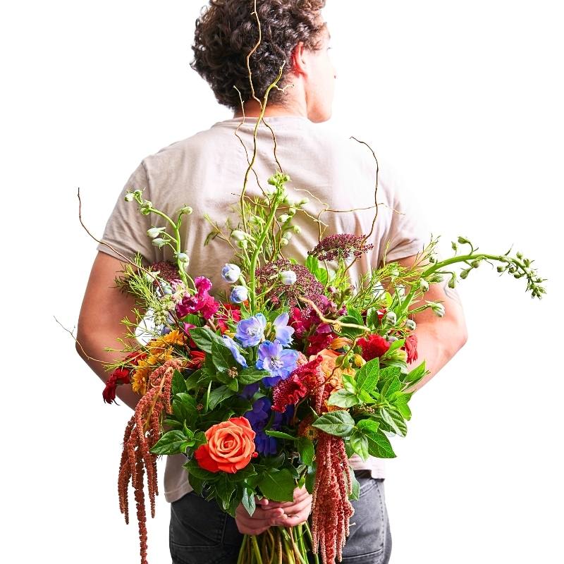 Wild and Free bouquet held behind back — bold orange rose, blue blooms, rich reds and wild greenery with trailing amaranthus