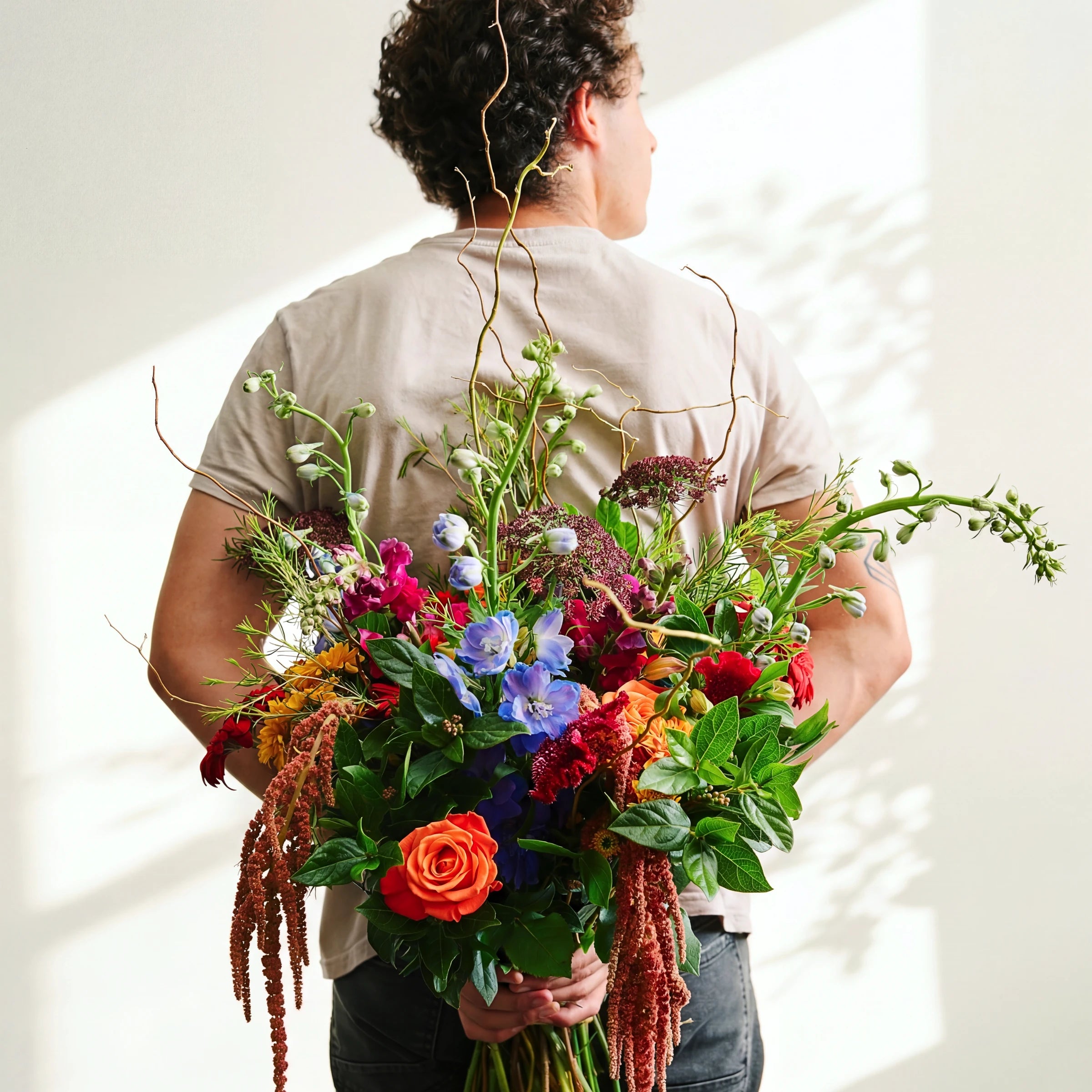 Wild & Free bouquet of orange roses, blue delphinium and trailing amaranthus with twisted willow, held in the sun