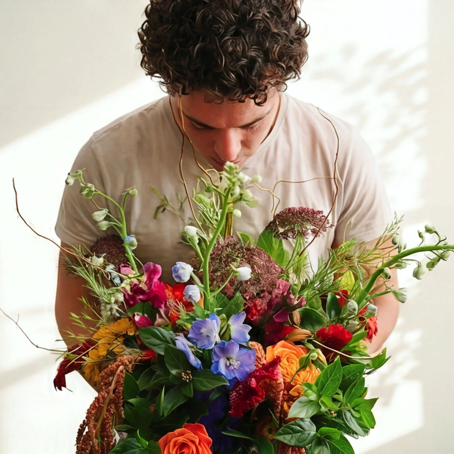 A man with curly dark hair wearing a beige t-shirt leans down to smell a large, vibrant, multicolored wildflower bouquet held in front of him against a bright, sunlit white background.