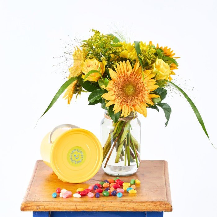 Bouquet of yellow flowers in a glass vase on a wooden table with a yellow container and colorful candies.