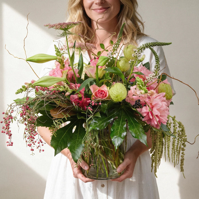 Smiling woman in a white linen dress holding pink roses, dahlias and lily buds in a clear glass vase, Fabulous Flowers South Africa