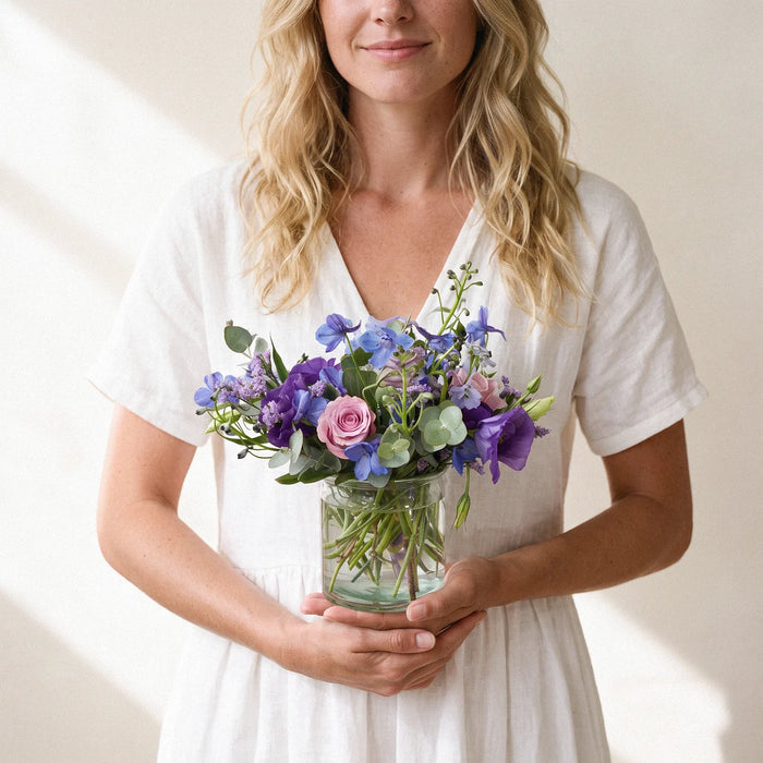 Bubbles and Blue, blue delphinium, purple lisianthus and lilac roses in a small glass vase, held in both hands by a smiling woman, Fabulous Flowers South Africa