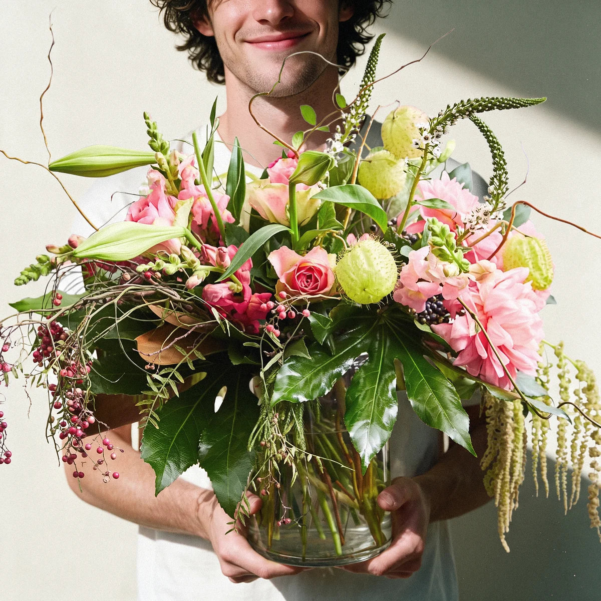 Smiling man in a white t-shirt carrying a glass vase of pink roses, dahlias and trailing amaranthus to give, Fabulous Flowers South Africa