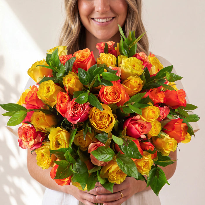 A smiling woman holds a large, vibrant bouquet of hybrid roses in shades of orange, yellow, and red, accented by lush green leaves, against a soft beige background.