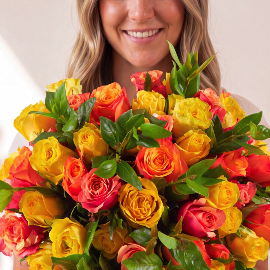 A smiling woman holding a large, vibrant bouquet of red, orange, and yellow roses with green leaves against a plain light background.