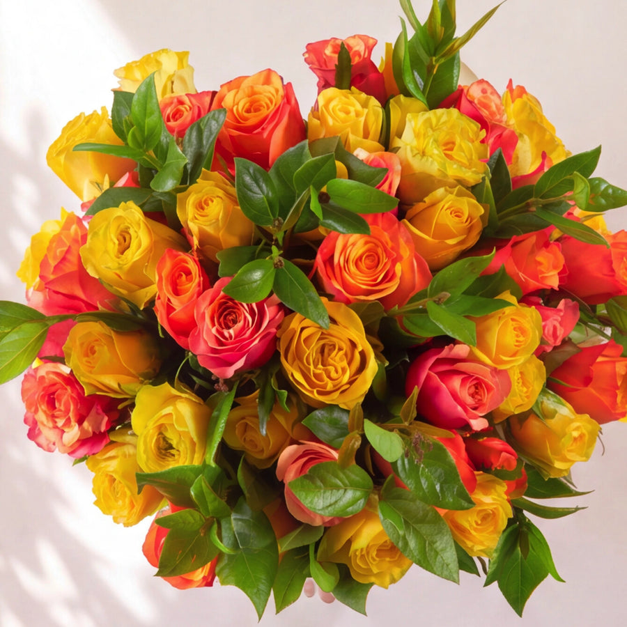 A dense bouquet of hybrid tea roses in vibrant yellow, orange, and red tones, interspersed with fresh green leaves, photographed from a top-down perspective against a clean white background.