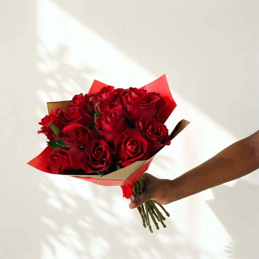 A dozen red roses hand-tied in red and gold wrap, held out by a florist from the Amour bouquet range