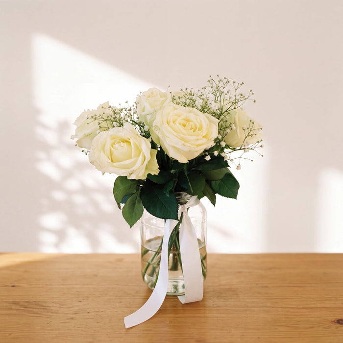 Angel White arrangement of ivory white roses and baby's breath in a glass jar with a white ribbon on a wooden table