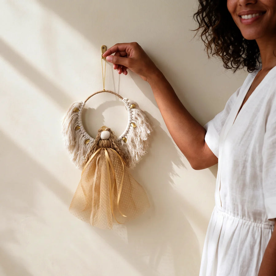 Woman in a white linen dress hanging a gold and cream macramé Christmas angel on a wall hook, Fabulous Flowers South Africa