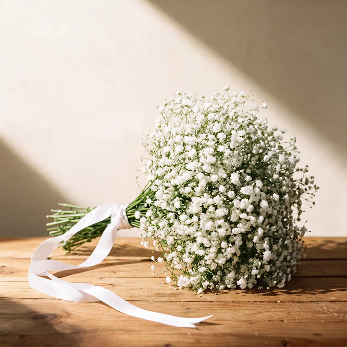 Hand-tied bouquet of white baby's breath with a trailing white satin ribbon lying on a pine table against a sunlit cream wall, Fabulous Flowers South Africa