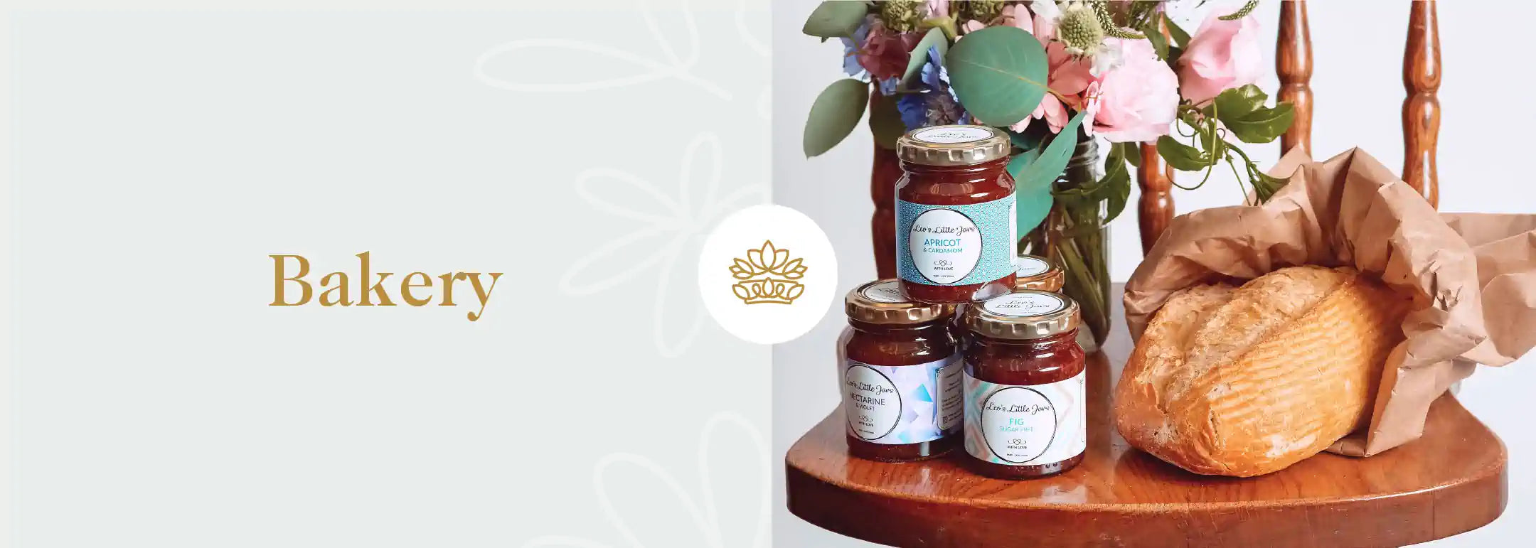 A rustic wooden table presents a selection of gourmet jams alongside a fresh, crusty loaf of bread, beside a vase of delicate flowers, evoking the homely charm of a 'Bakery' with a flower crown emblem - Fabulous Flowers and Gifts.