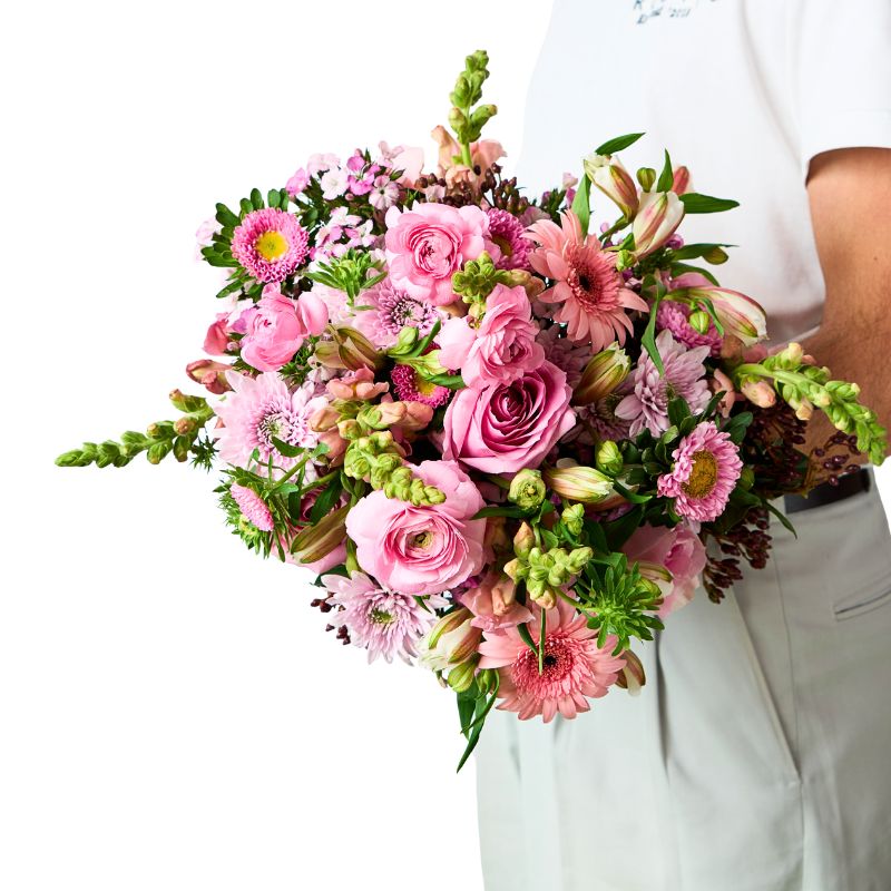 Florist in white holding a large hand-tied bouquet of pink roses, ranunculus, gerberas, snapdragons, alstroemeria and greenery against a white background.