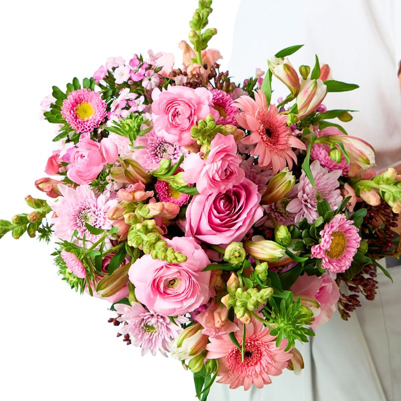 Close-up of a lush pink bouquet—roses, ranunculus, gerberas, chrysanthemums, alstroemeria and fresh greenery—cradled by a florist on a bright white backdrop.