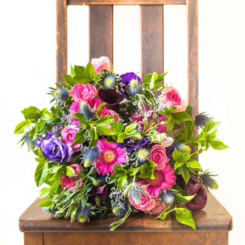 Vibrant hand-tied bouquet of pink roses and gerberas with purple lisianthus, plum calla lilies, blue thistle and lush greenery on a rustic wooden chair.”
