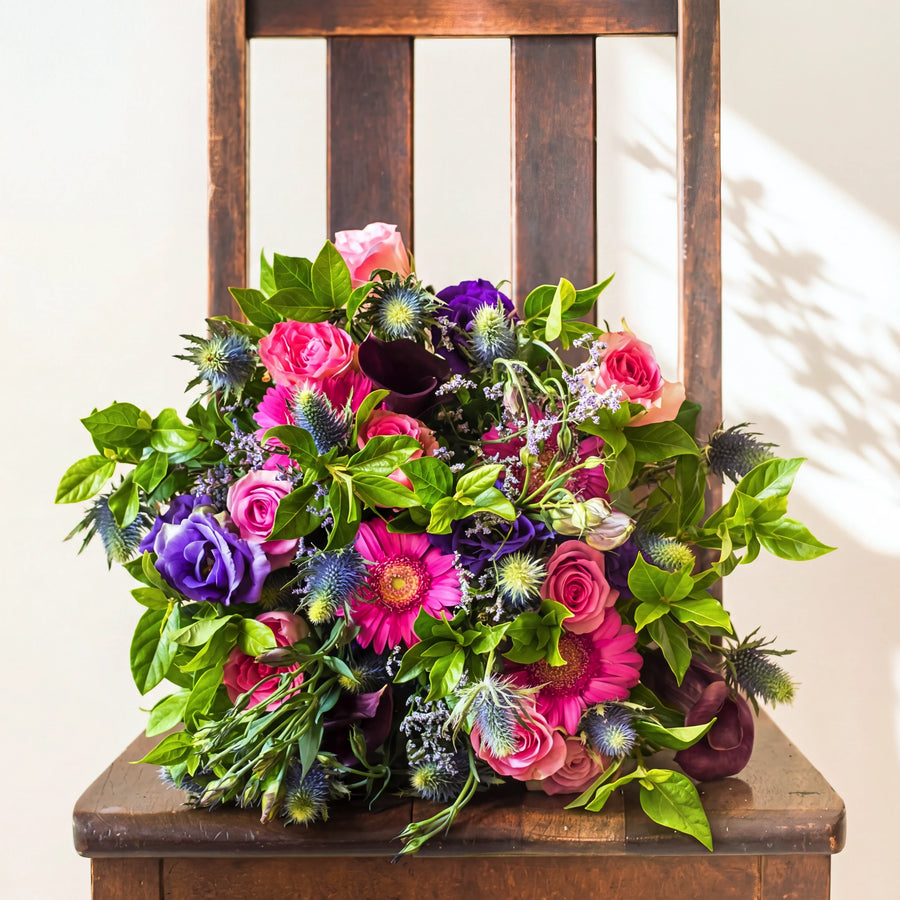 Berry Bloom bouquet of pink roses, hot pink gerberas, purple lisianthus and blue thistle on a wooden chair